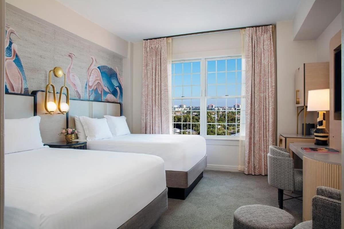 A hotel room with two beds, a desk, a chair, a window with curtains, and a wall with a painting.