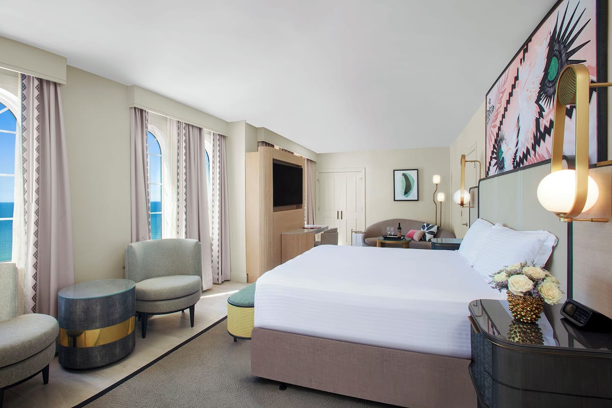 A hotel room with a large bed, two armchairs, a small side table, a mirror, and a painting on the wall.