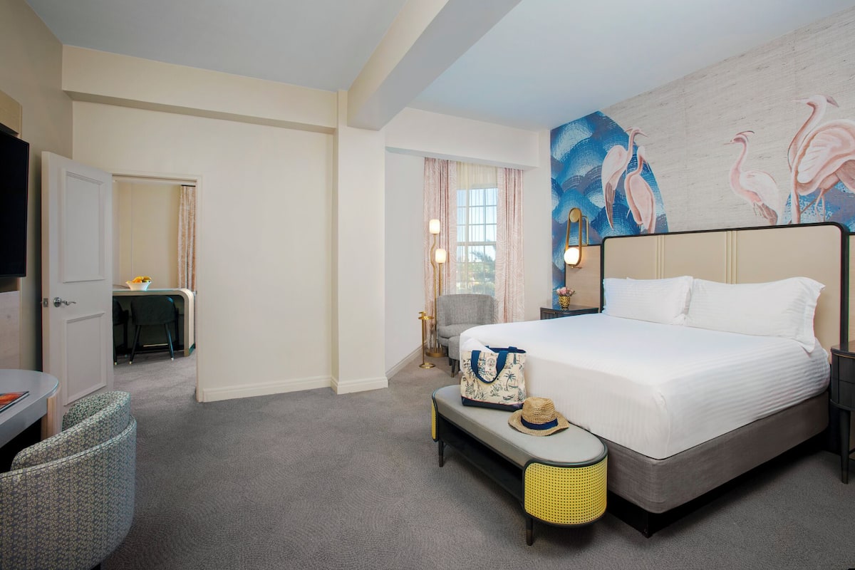A hotel room with a large bed, a desk with a chair, a small table, a bench, and a wall with a painting.