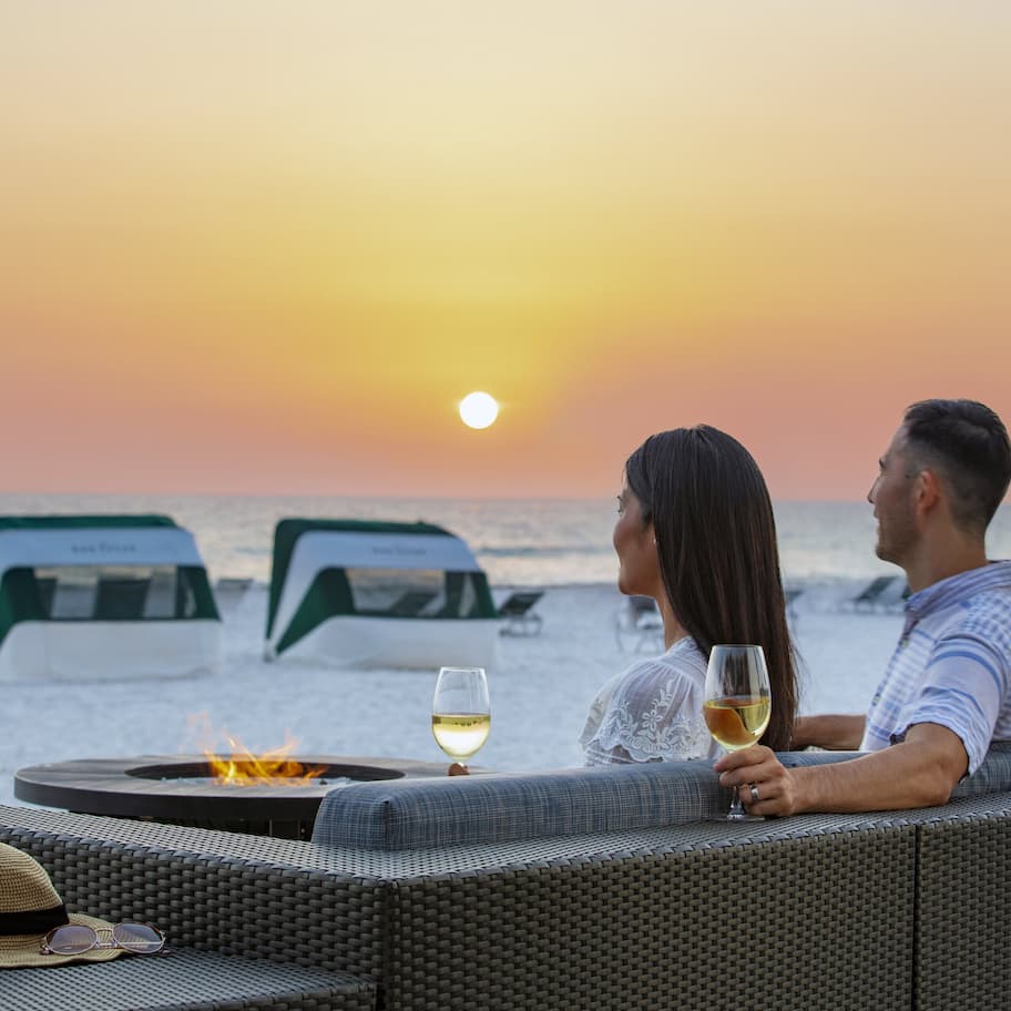 A couple sitting on a wicker sofa overlooking the ocean at sunset, with a fire pit and glasses of wine on the table.