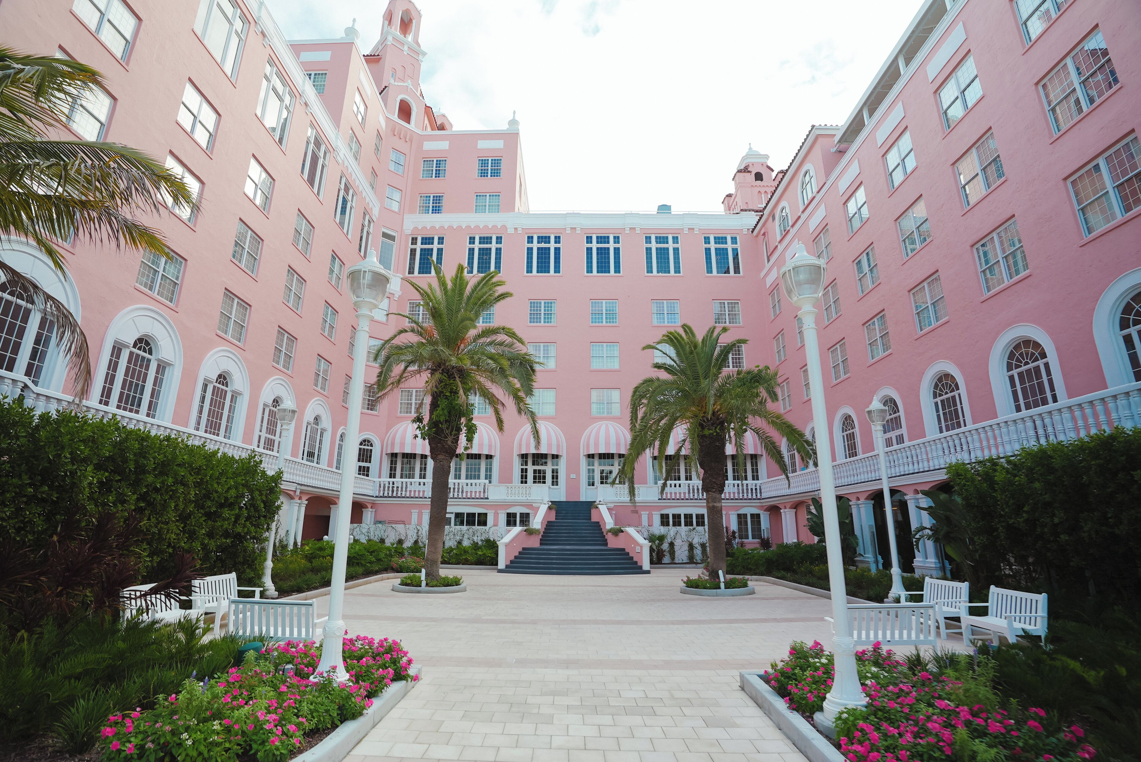 A pink building with a central staircase, surrounded by palm trees and benches.