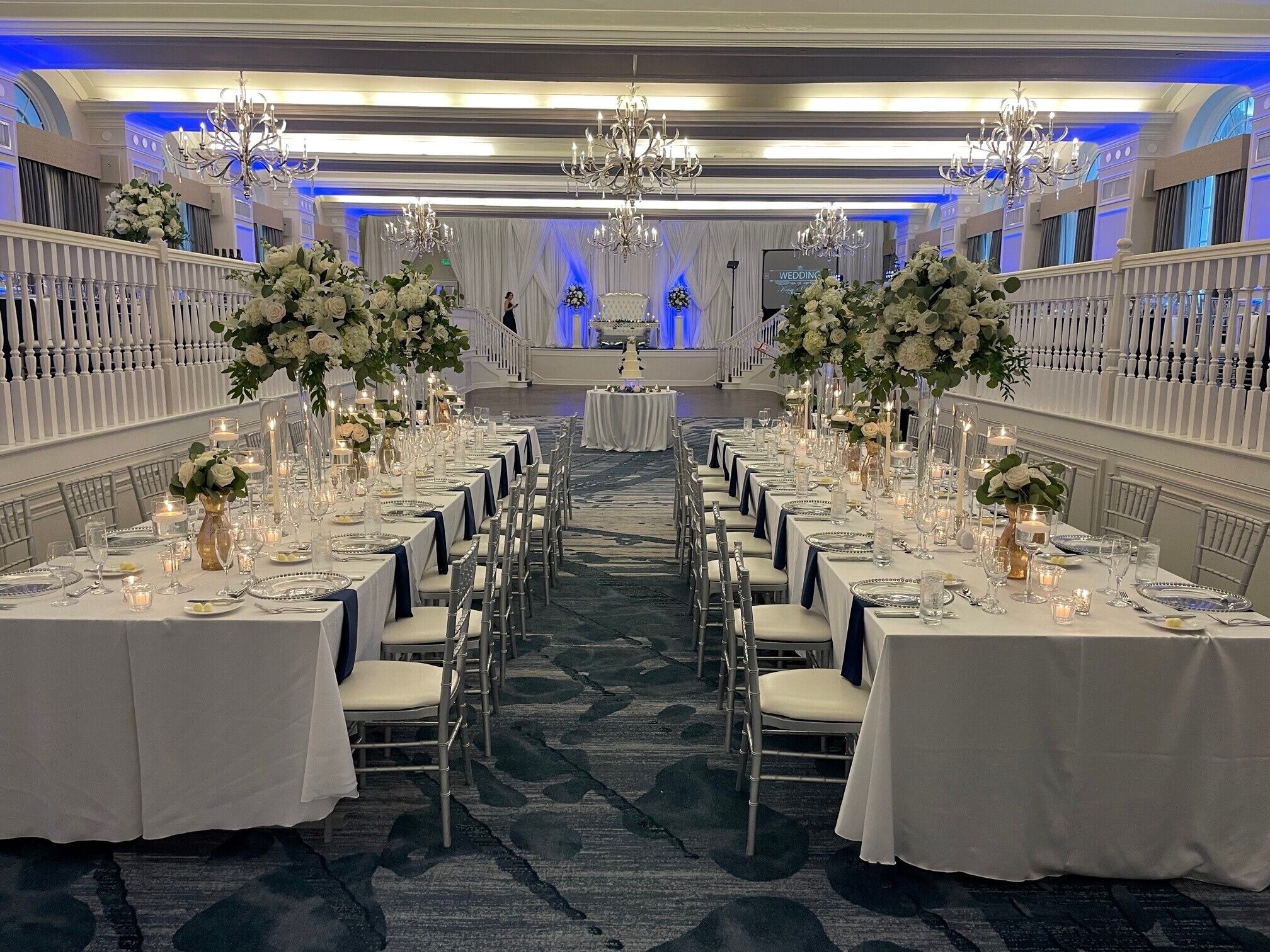 A banquet hall with rows of tables set for a wedding reception, adorned with white flowers and candles.
