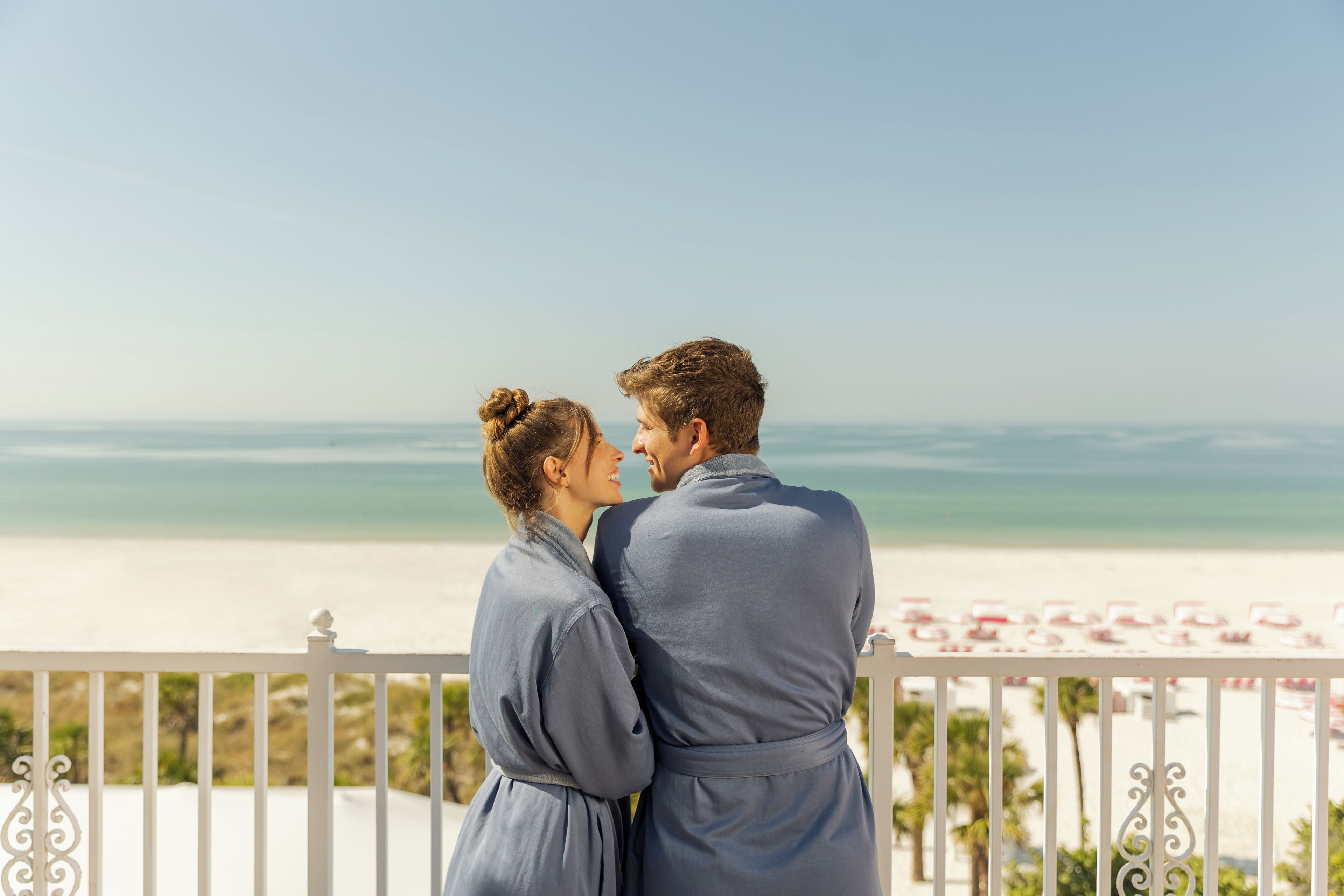 Una pareja parada en un balcón con vista a una playa, donde se distinguen claramente el océano y sombrillas.