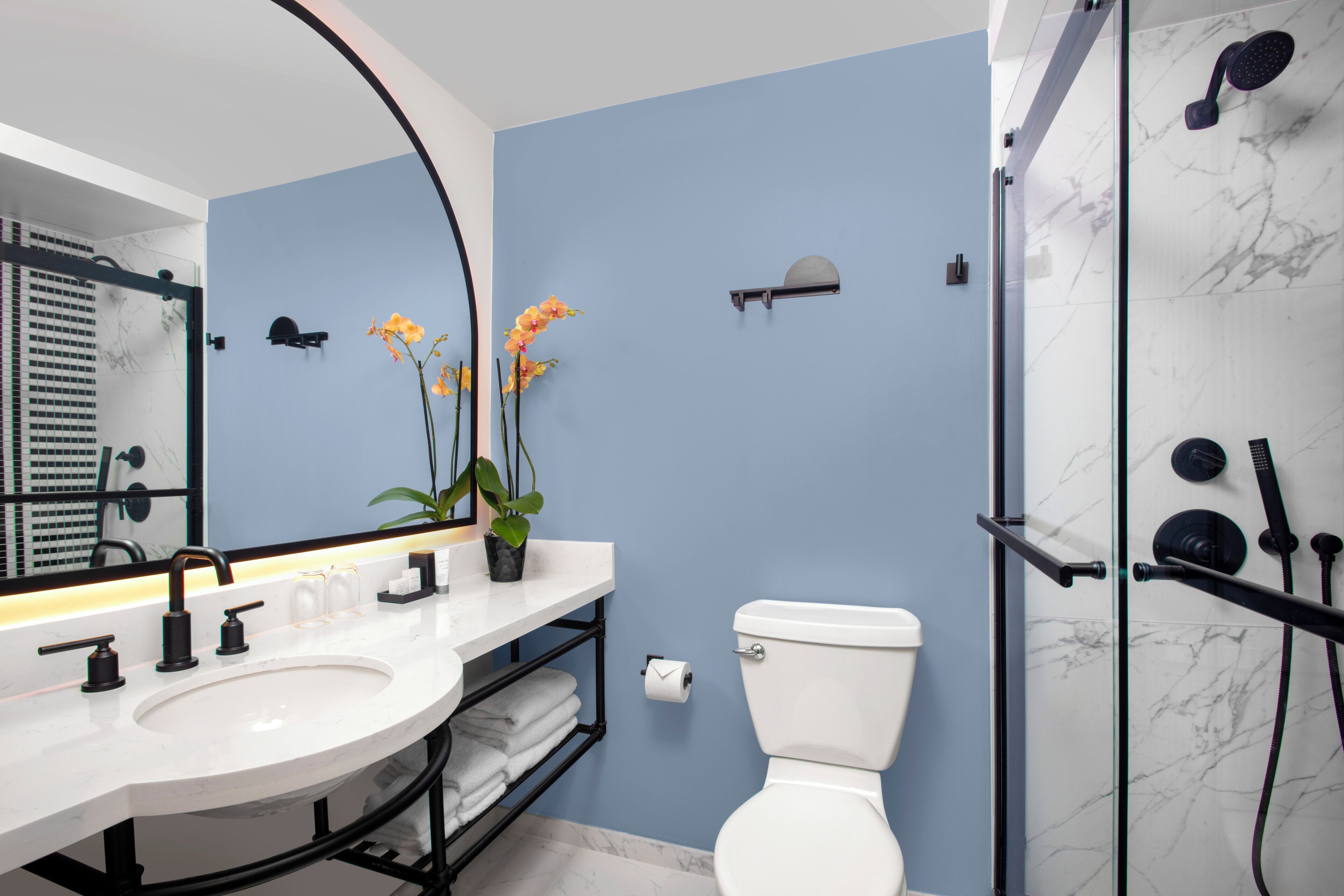 A modern bathroom with a white sink, two black faucets, a white toilet, and a glass-enclosed shower with a marble wall.