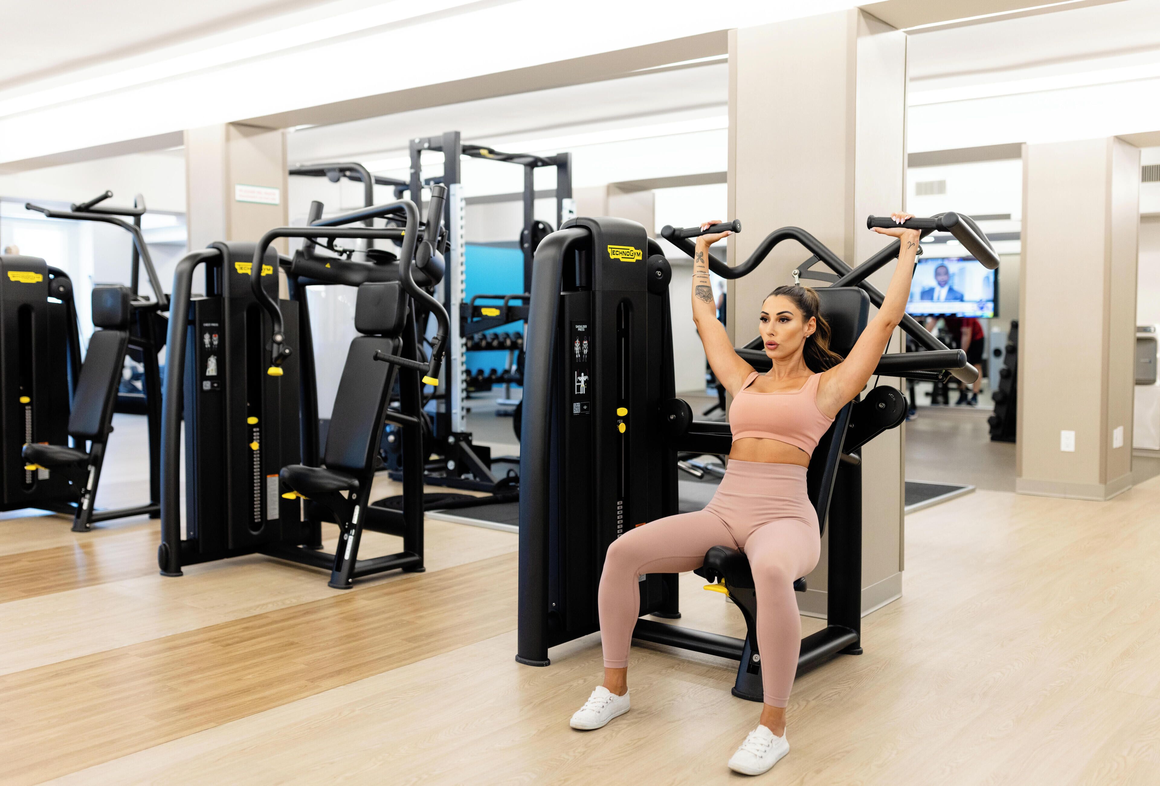 Eine Frau trainiert auf einem Gerät in einem Fitnessstudio, im Hintergrund sind verschiedene Geräte und ein Fernseher zu sehen.