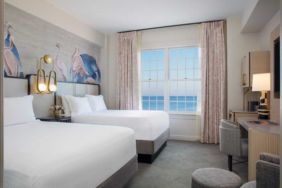 A hotel room with two beds, a desk, a chair, and a view of the sea.