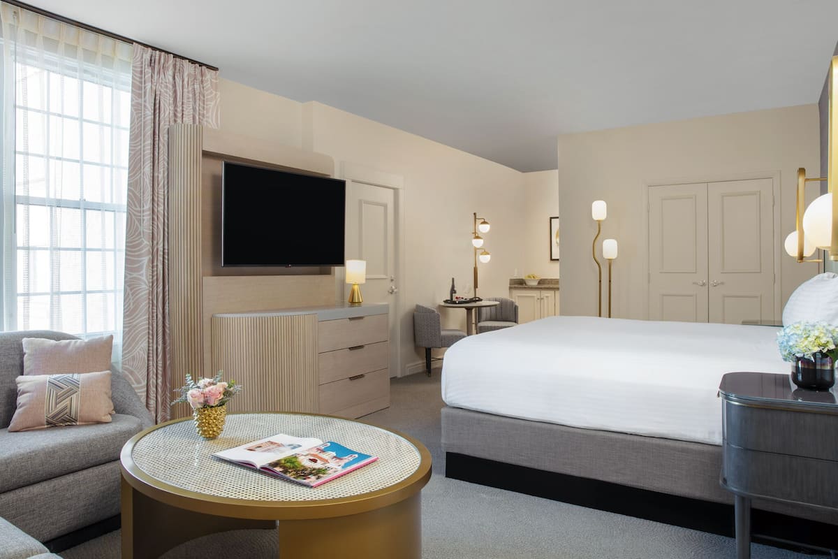 A modern hotel room with a large bed, a sofa, a round table with a magazine, a TV mounted on the wall, and a desk with a lamp.