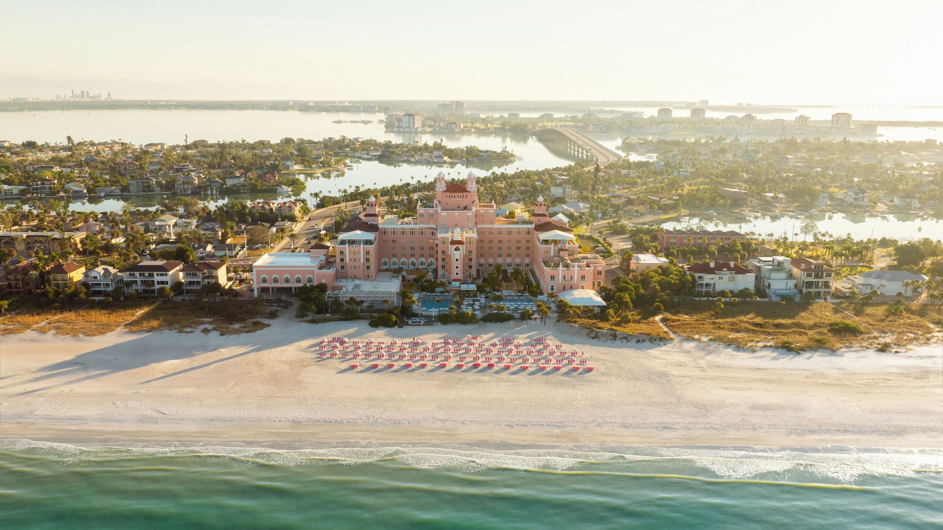 A large resort with a central building surrounded by water, a beach with umbrellas, and a marina.