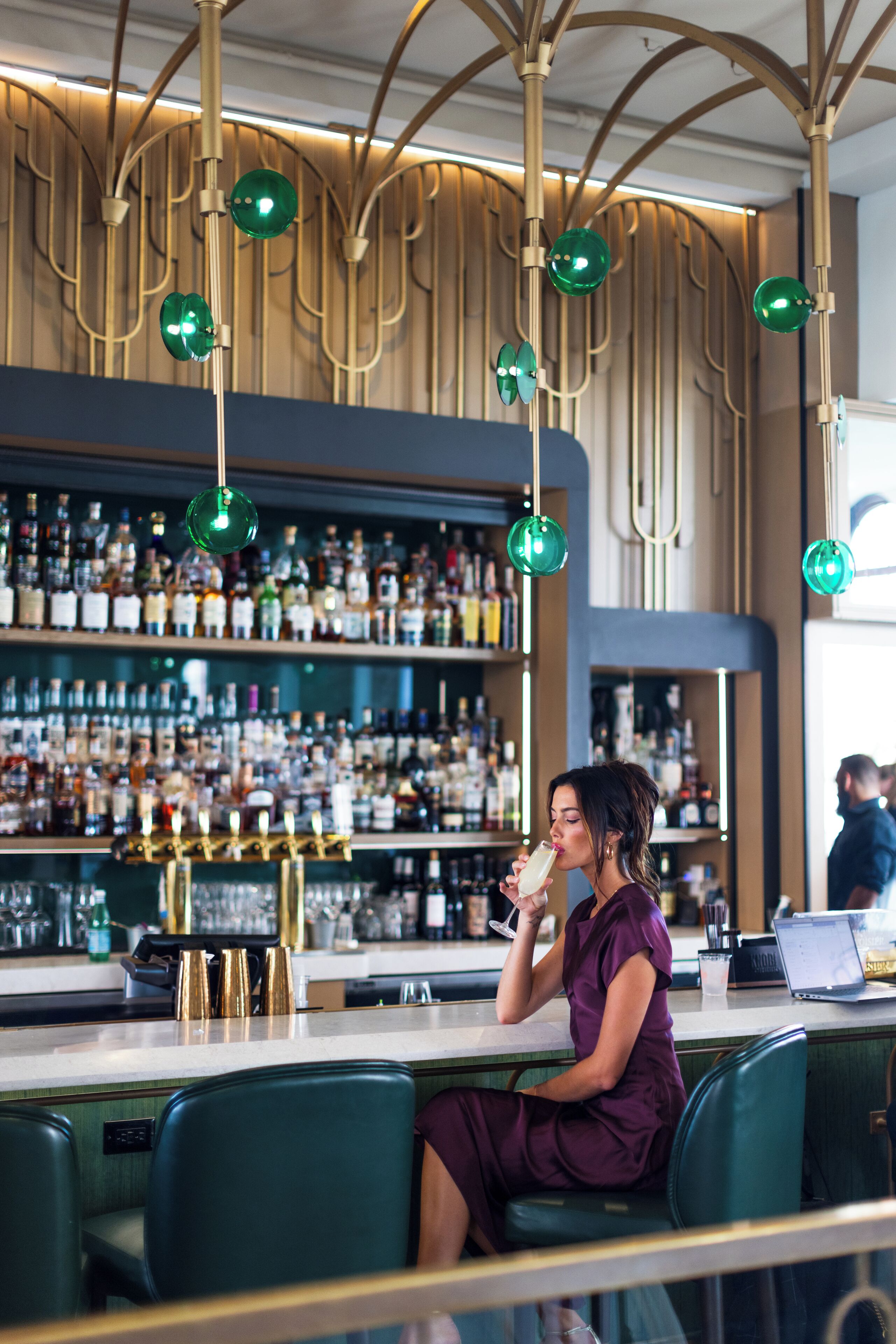 Una zona de bar con una mujer sentada en la barra, una estantería repleta de licores al fondo y una lámpara moderna.
