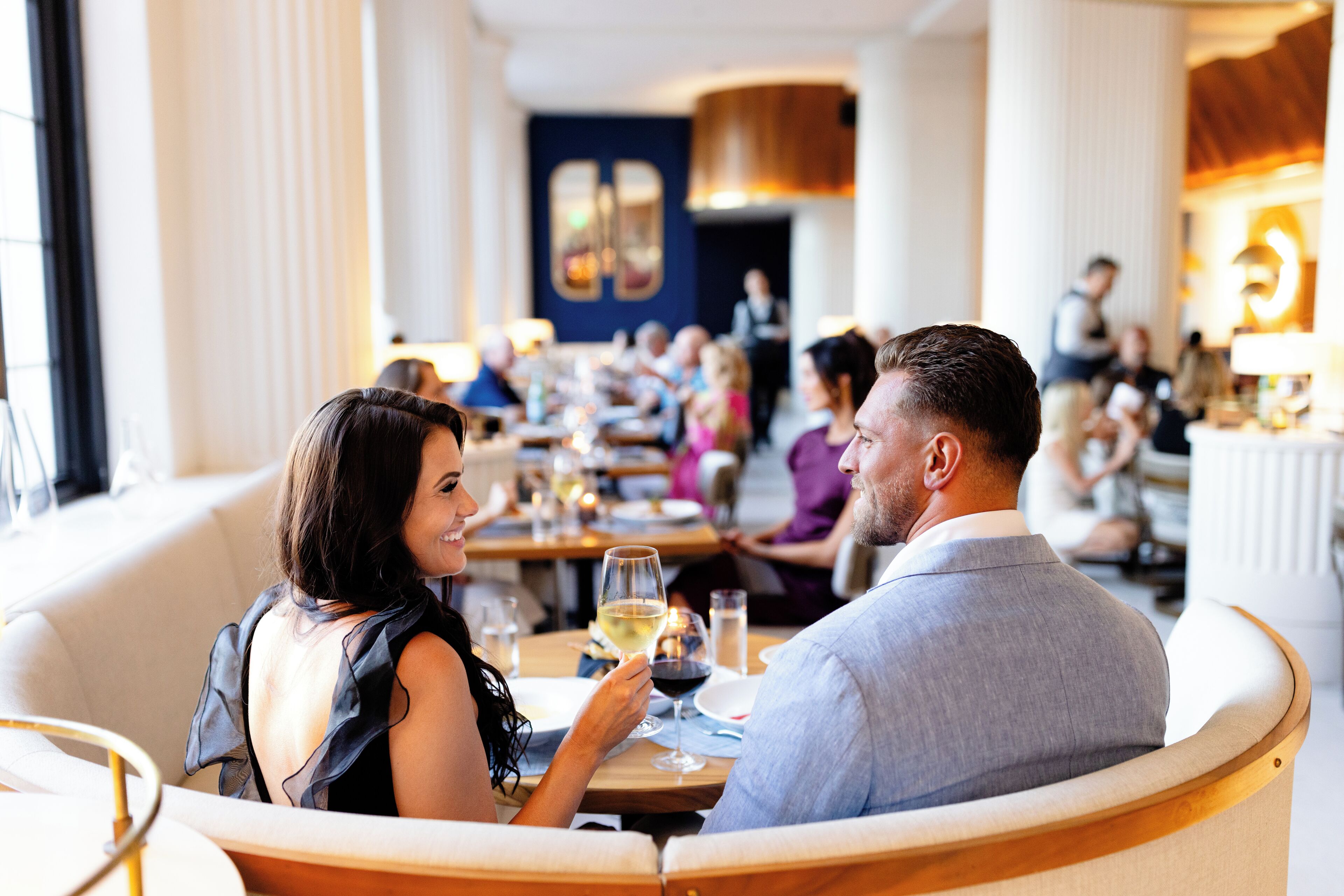 A couple dining in a modern restaurant with a view of other diners and a waiter in the background.
