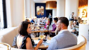 A couple dining in a modern restaurant with a view of other diners and a waiter in the background.