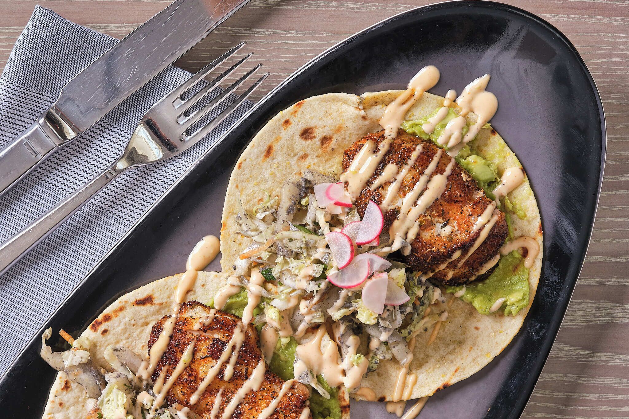 Un plato con dos tacos, cada uno con una hamburguesa a la parrilla, aguacate y salsa cremosa, acompañado de un tenedor y cuchillo sobre una servilleta.