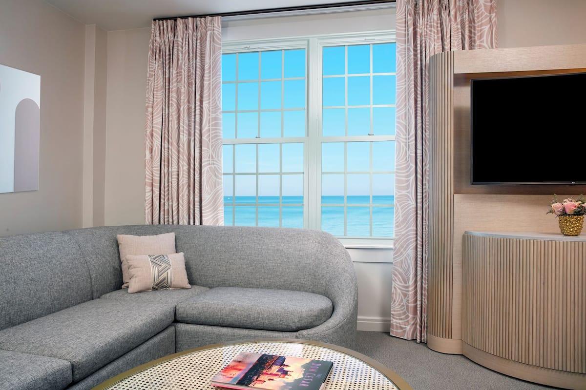 A modern living room with a grey sofa, a round coffee table with a book, a flat-screen TV mounted on a wooden panel, and a window with a view of the sea.