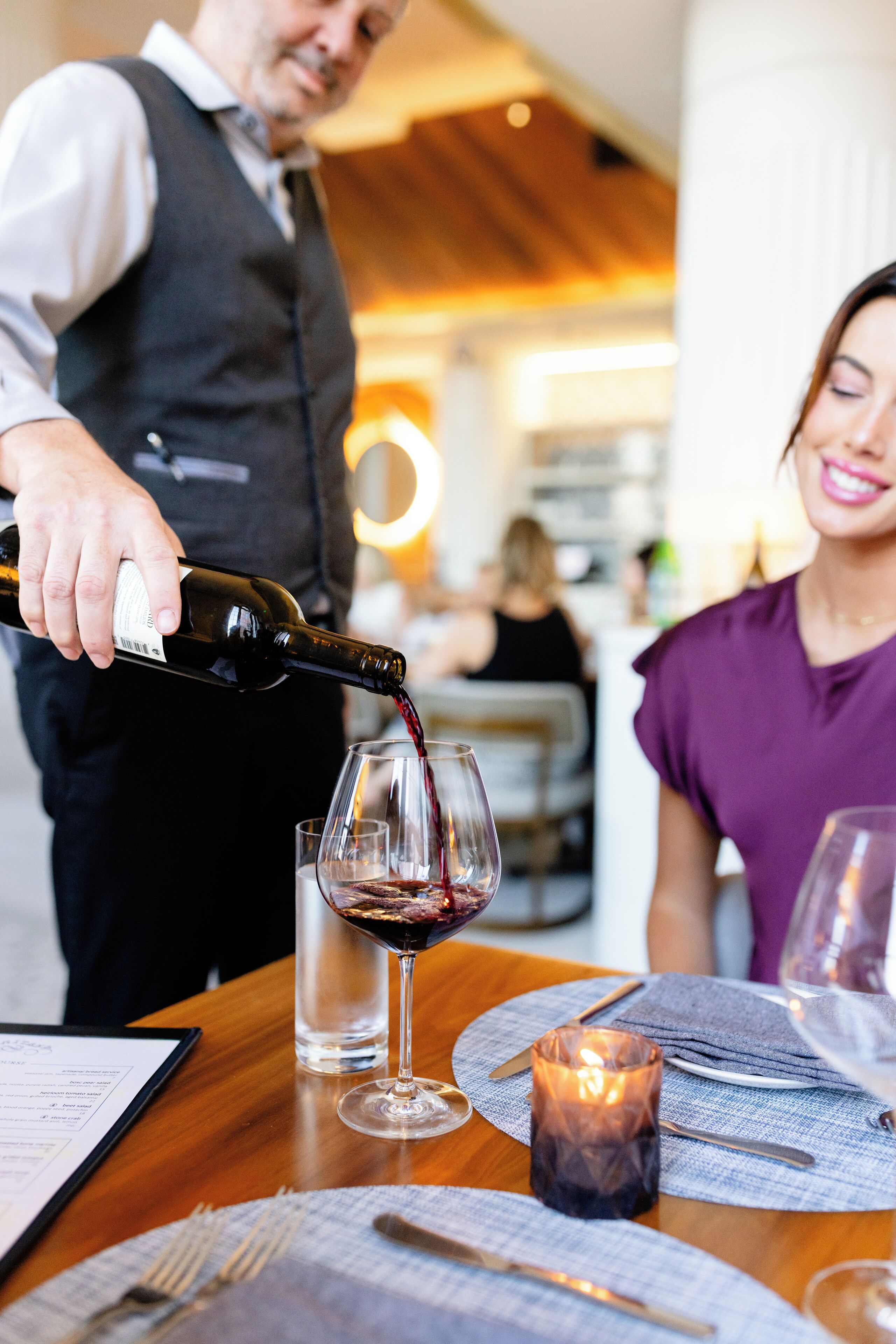 Un serveur verse du vin dans un verre sur une table de restaurant, où se trouvent une bougie et un menu.