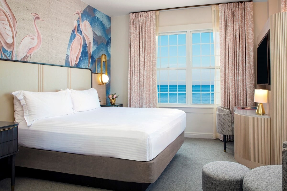 A hotel room with a large bed, a view of the sea, a painting of birds on the wall, and a TV on the wall.