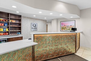 Zona de recepción de hotel con mostrador blanco, un letrero de 'Quality by Choice Hotels', un televisor montado en la pared y estantes con aperitivos.