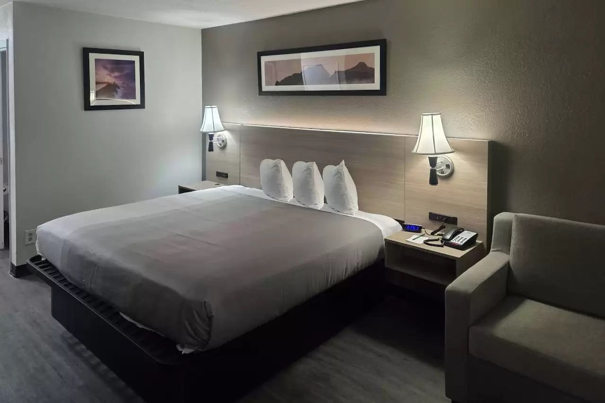 A hotel room with a large bed, two bedside lamps, a nightstand with a phone, and a chair.
