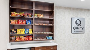 Un ripiano pieno di snack assortiti, tra cui patatine e altri prodotti, un frigorifero con bevande e un cartello che indica ‘Qualità by Choice Hotels’.