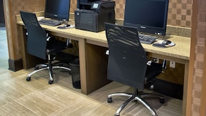 Un espacio de trabajo con dos escritorios, cada uno con un monitor de computadora, teclado y ratón. Hay dos impresoras sobre los escritorios, y la habitación tiene suelo de baldosas y paneles de madera en las paredes.