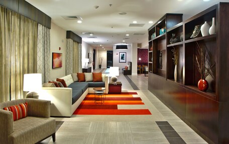 A modern hotel lobby with a sofa, armchairs, a dining area, and a bookshelf with decorative items.