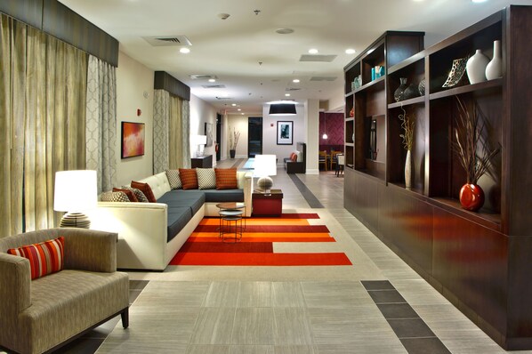 A modern hotel lobby with a sofa, armchairs, a dining area, and a bookshelf with decorative items. - Holiday Inn Charlotte Airport by IHG (Charlotte)