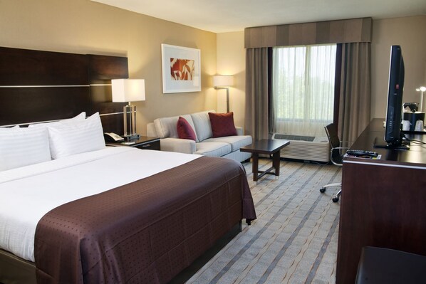 A hotel room with a large bed, a desk, a chair, a sofa, a TV, and a window with curtains. - Holiday Inn Charlotte Airport by IHG (Charlotte)