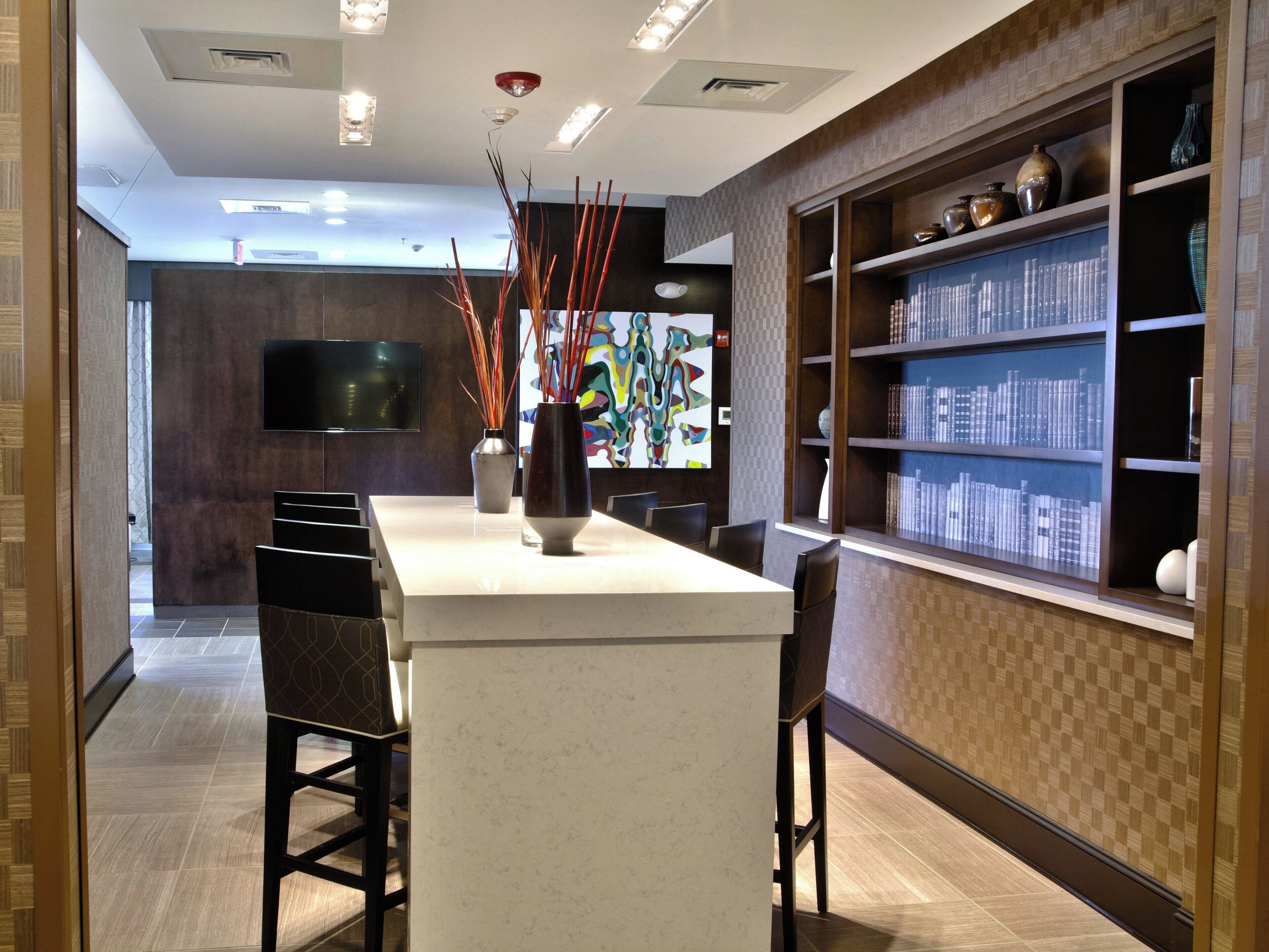 A modern kitchen with a central island, bar stools, a flat-screen TV, and a bookshelf with decorative items.