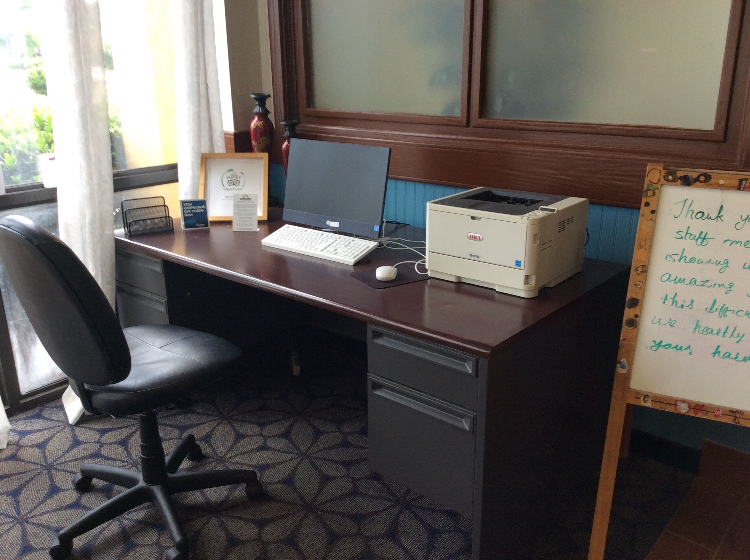 A desk with a laptop, a desktop computer, and a printer. There is a chair, a keyboard, a mouse, and a whiteboard with handwritten notes.