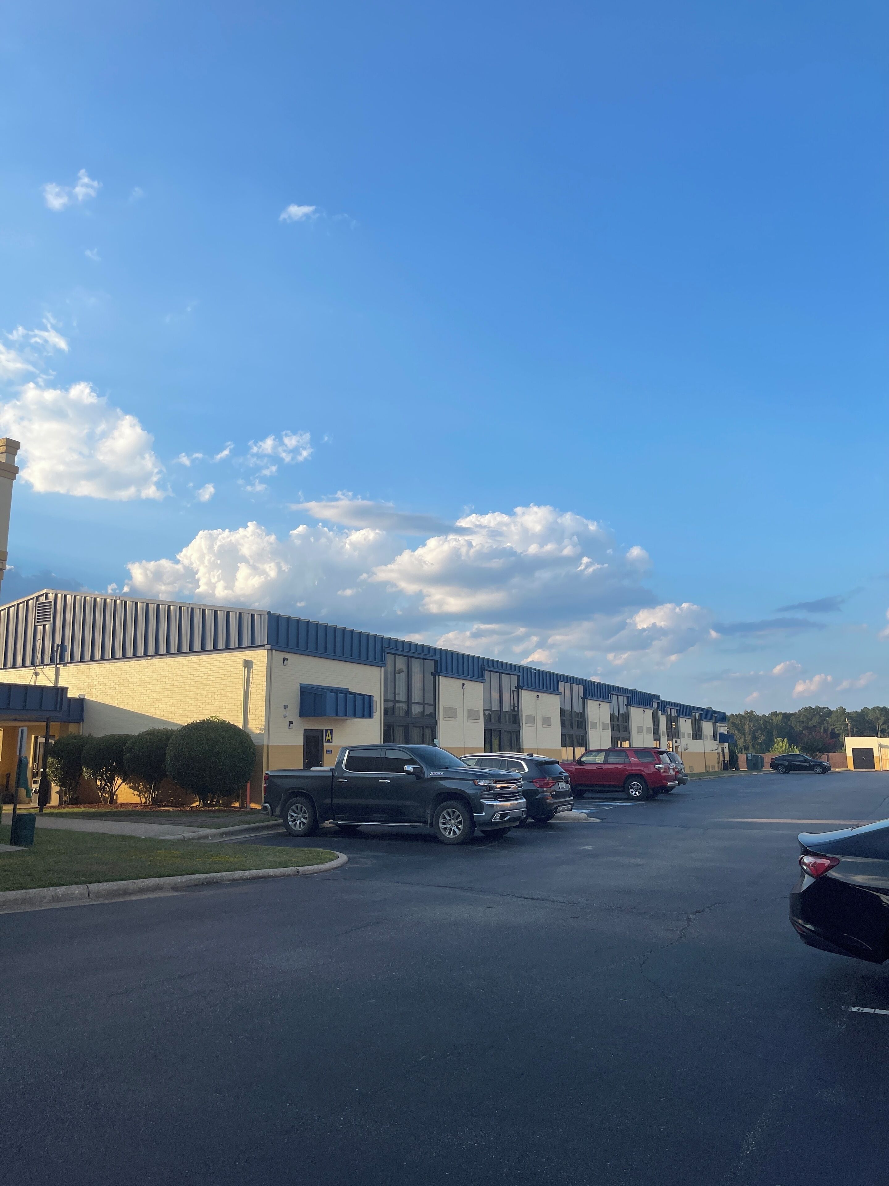 A parking lot with several cars, a building with a blue and white facade, and a clear sky with clouds.