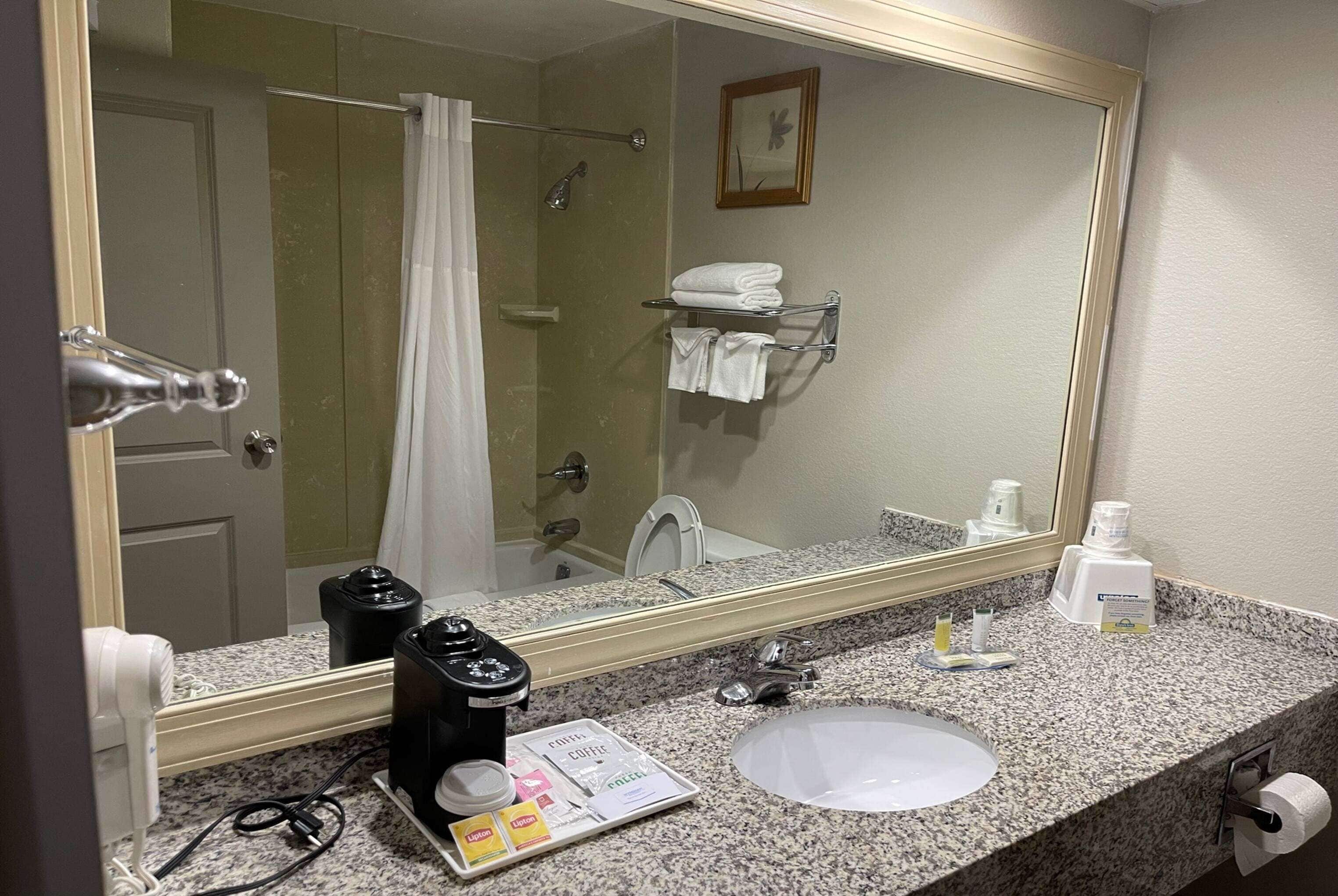 Baño de hotel con lavamanos, espejo, toallero, cortina de ducha y cafetera.