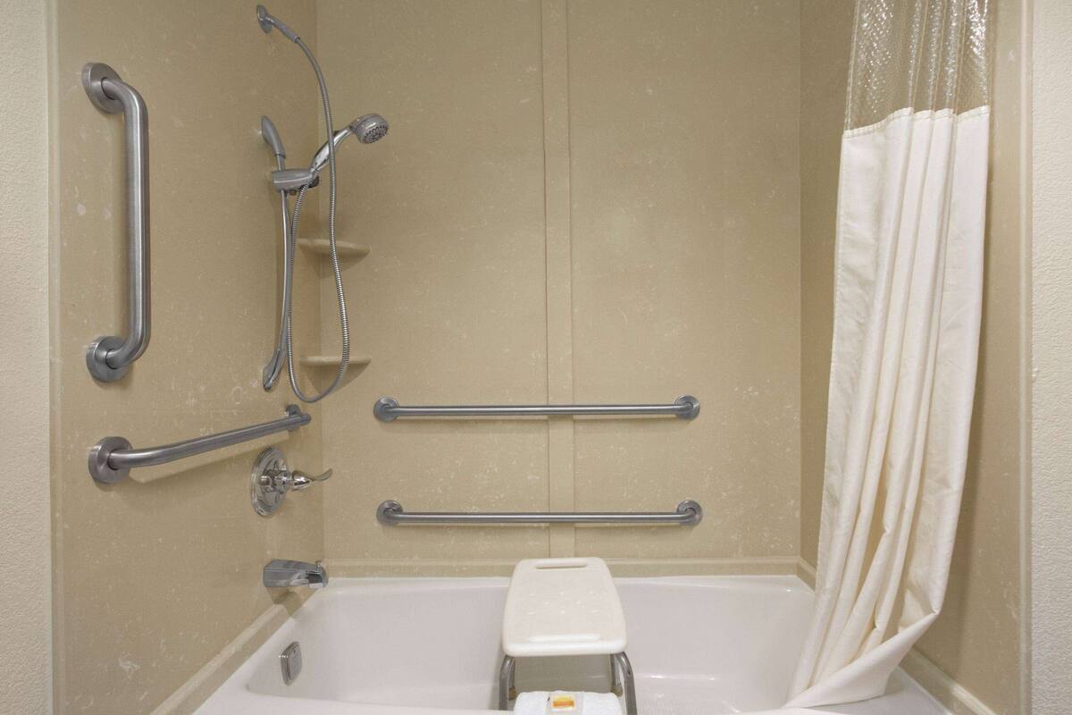Une salle de bain équipée d’une baignoire, de barres d’appui, d’un siège de douche et d’un rideau de douche blanc.
