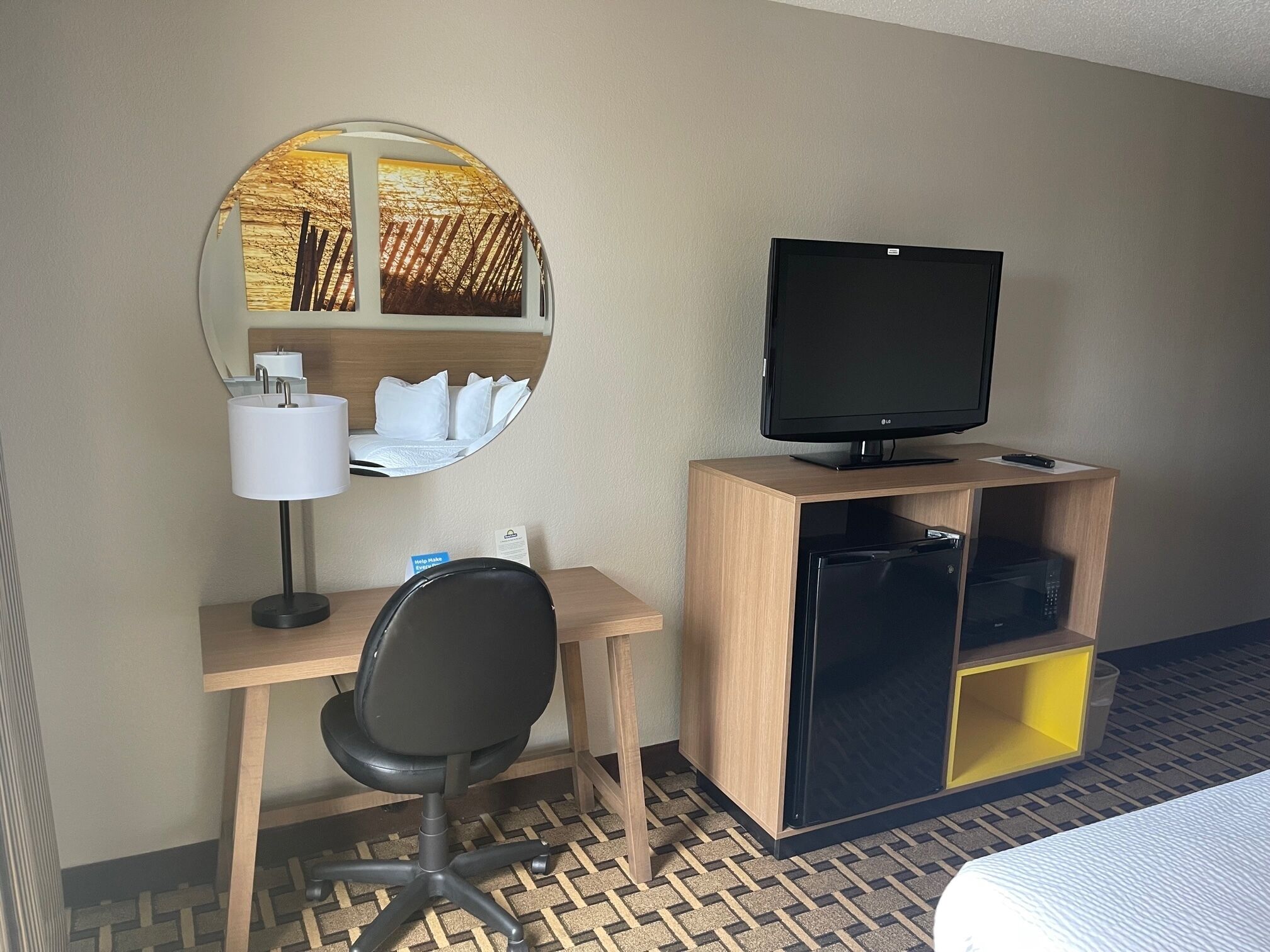 Ein Hotelzimmer mit einem Holztisch, einem schwarzen Bürostuhl, einem Fernseher auf einem hölzernen Standfuß, einem Spiegel mit Blick auf einen Sonnenuntergang und einem Bett mit weißer Tagesdecke.
