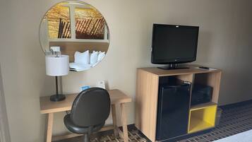 Habitación de hotel con escritorio de madera, silla de oficina negra, televisor sobre un mueble de madera, espejo con vista a un atardecer y cama con cubrecama blanco.