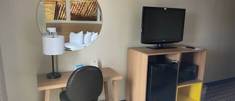 A hotel room with a wooden desk, a black office chair, a television on a wooden stand, a mirror with a view of a sunset, and a bed with a white comforter.