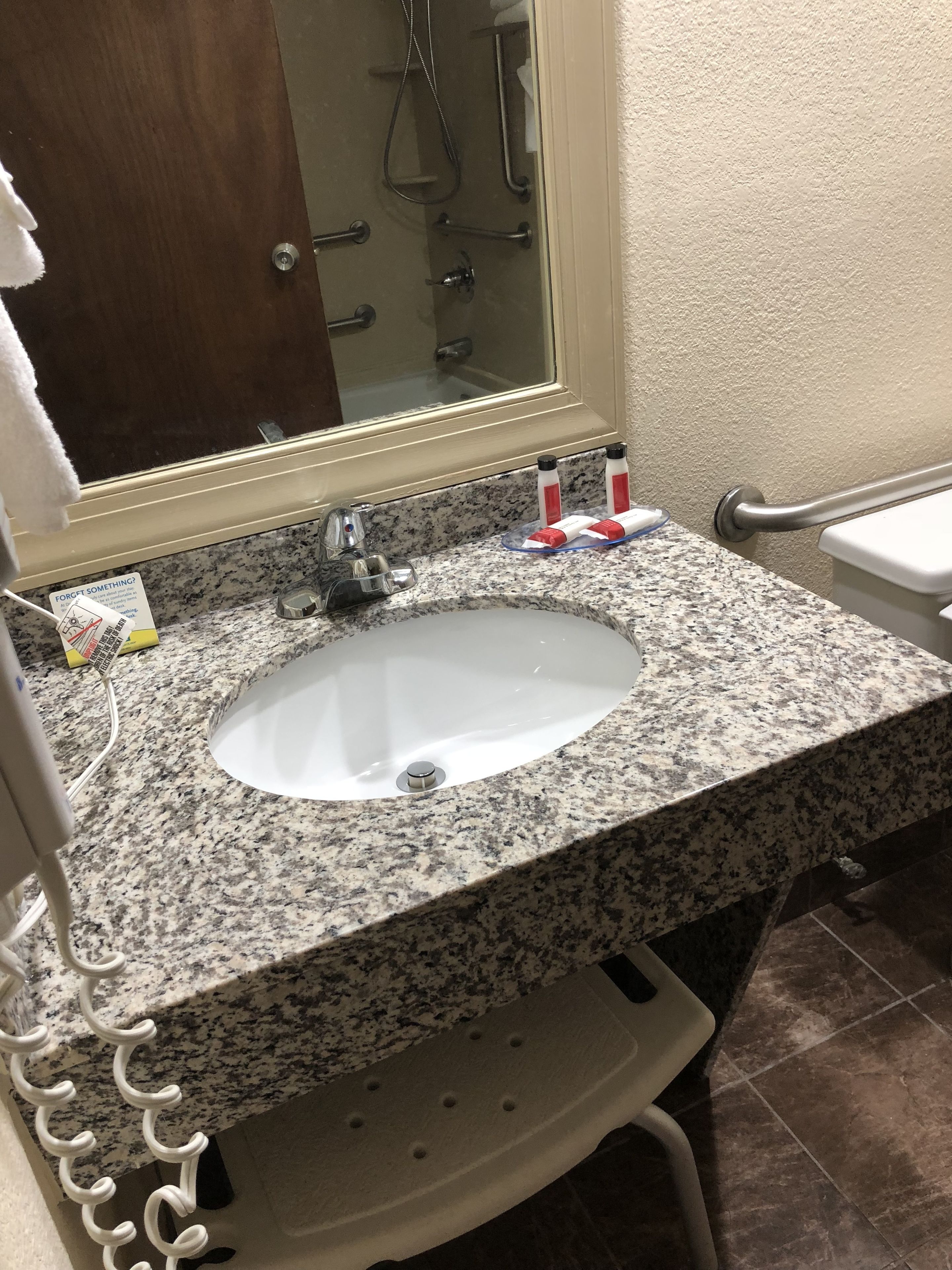 Un baño con encimera de granito, un lavabo blanco, un espejo y una zona de ducha visible a través de la puerta de cristal.