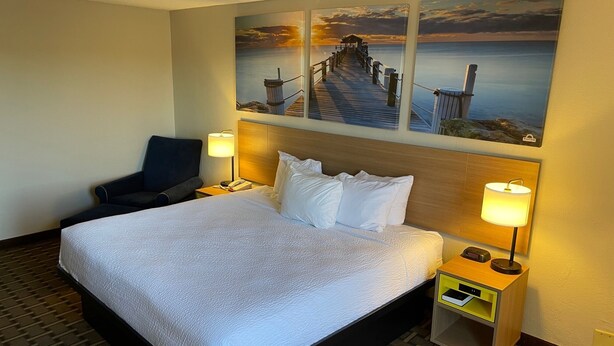 Habitación de hotel con una cama grande, dos mesitas de noche, una silla y una obra de arte que representa un muelle y un atardecer.
