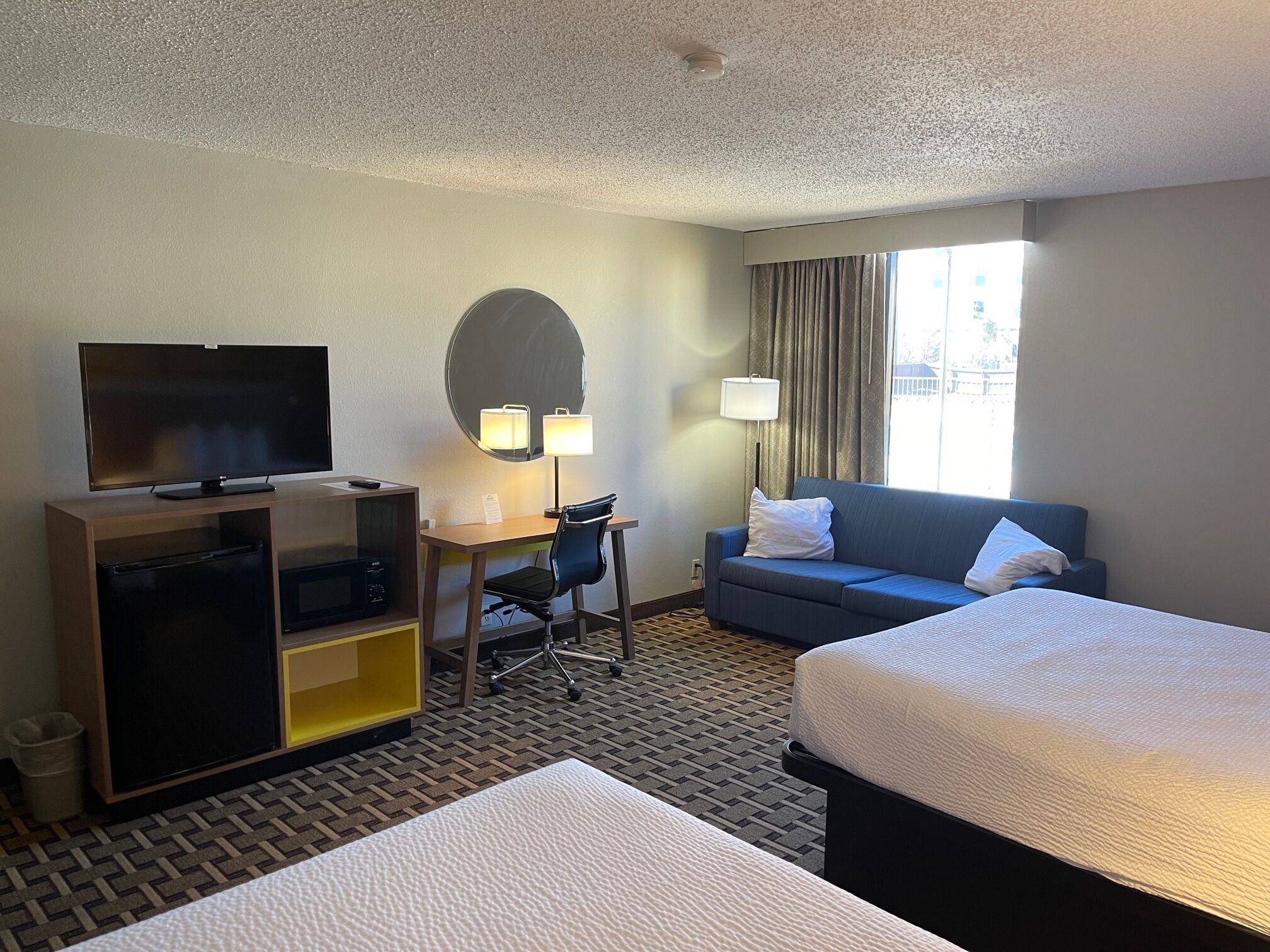 Une chambre d’hôtel comprenant un lit, un bureau avec une chaise, une télévision, un canapé et une fenêtre avec des rideaux.