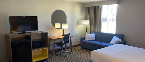 A hotel room with a bed, a desk with a chair, a television, a sofa, and a window with curtains.