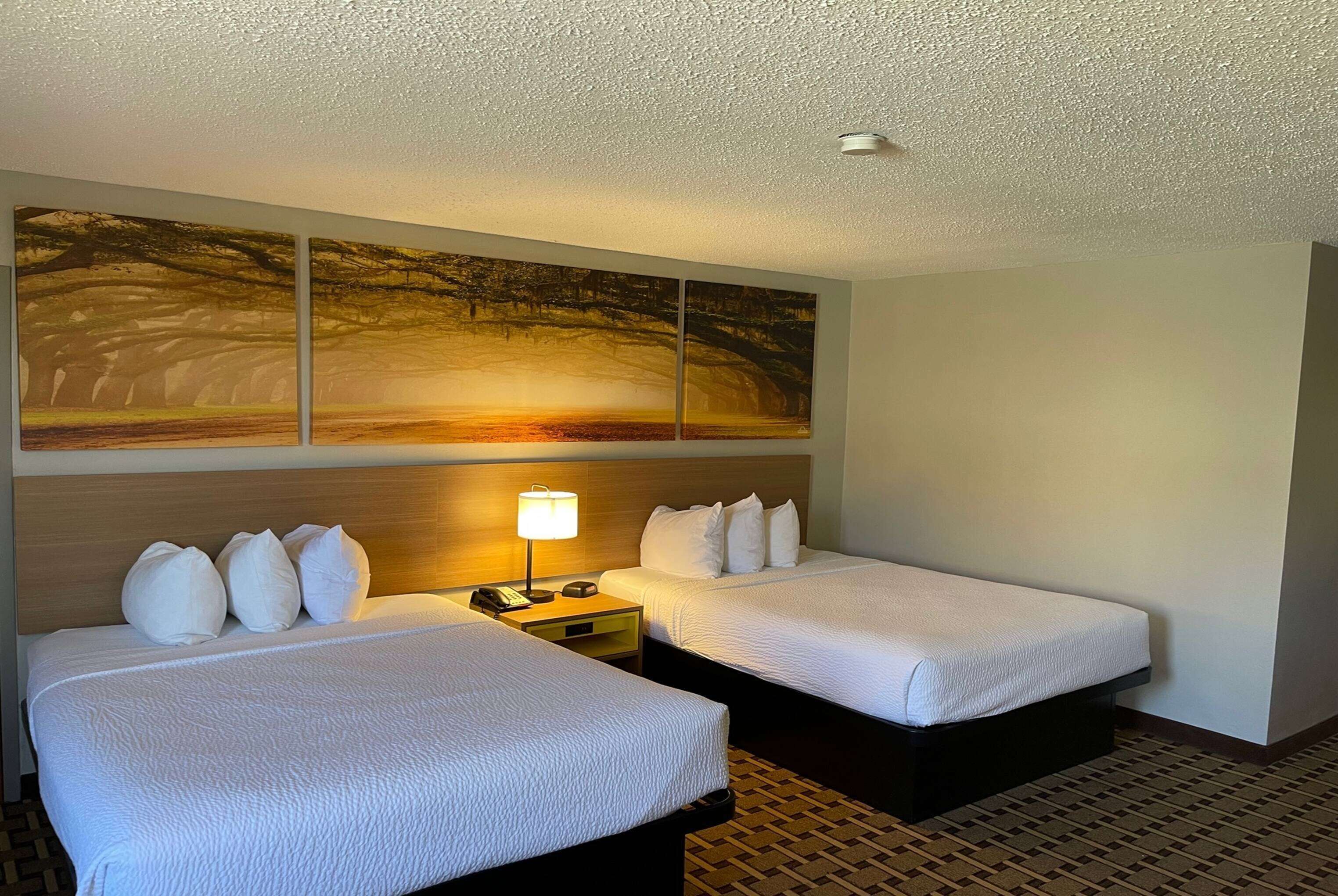A hotel room with two beds, a large wall art piece, a nightstand with a lamp, and a patterned floor.