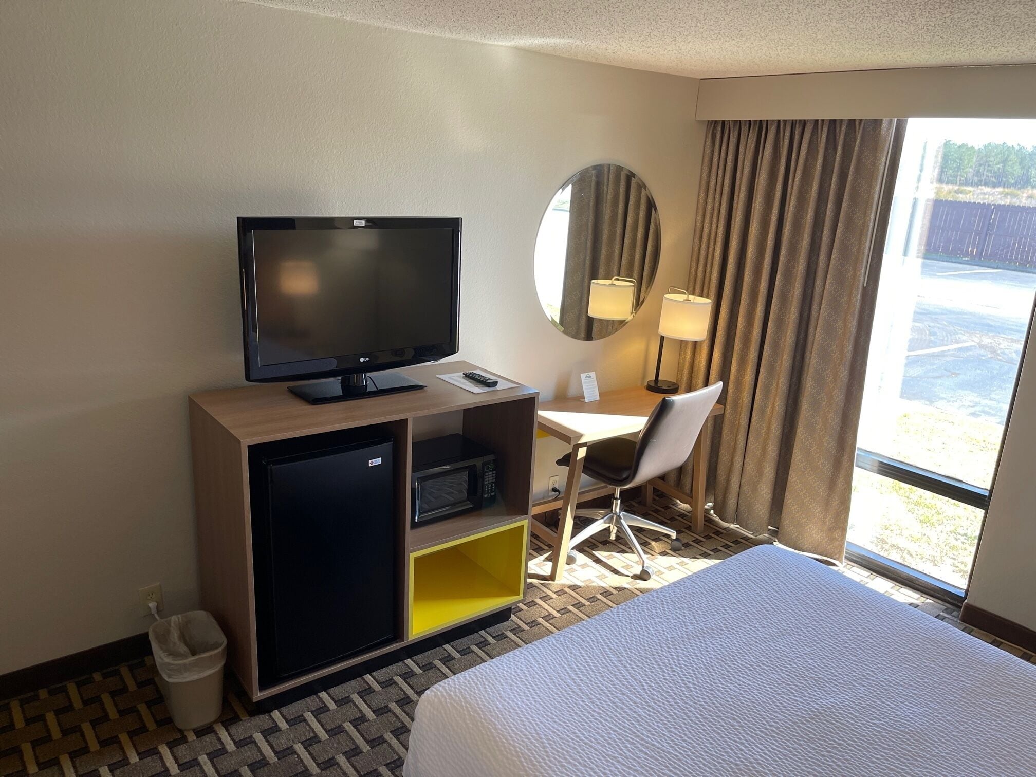 Habitación de hotel con cama, escritorio con televisor, microondas, basurero y ventana con cortinas.
