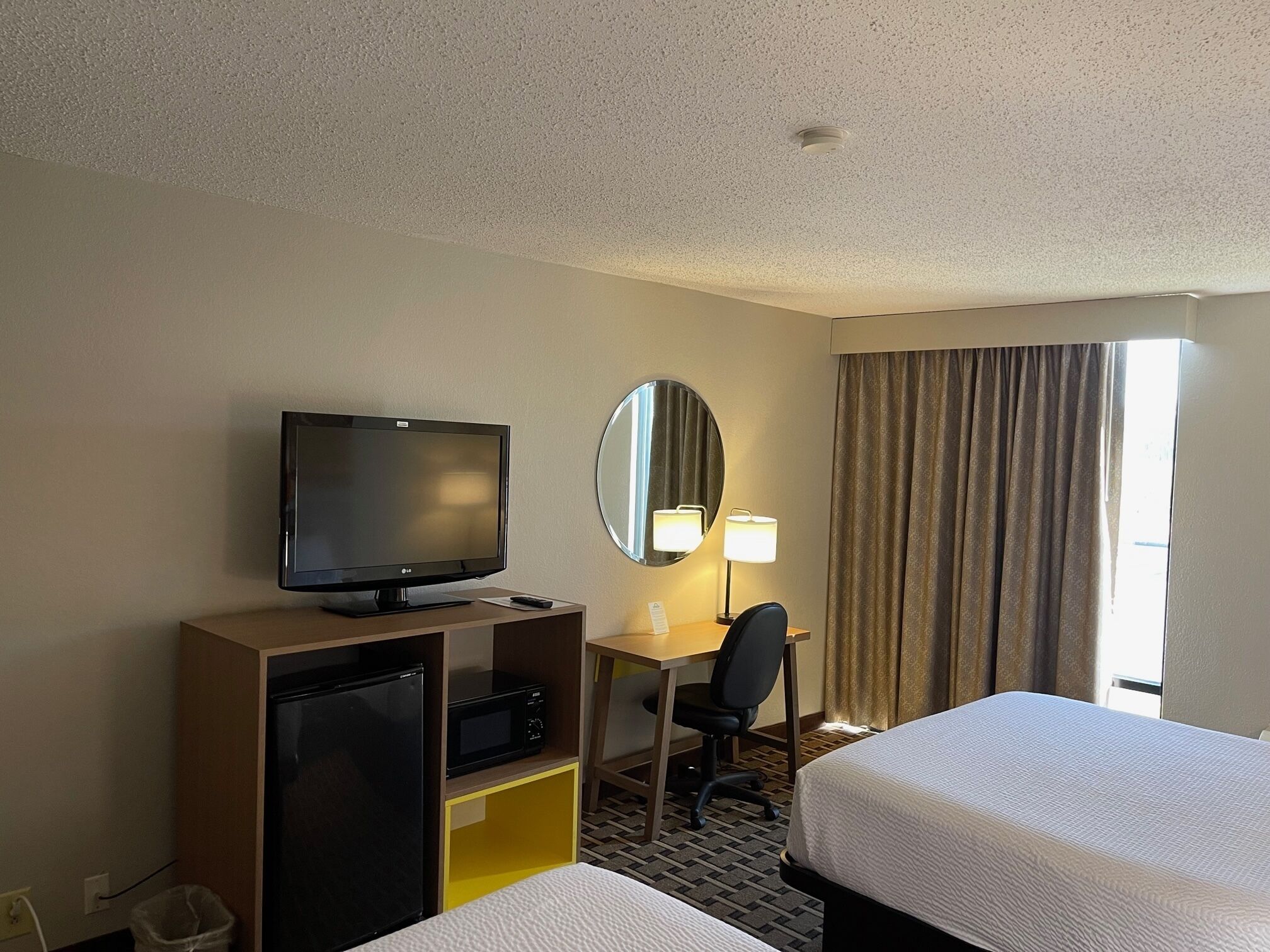 Habitación de hotel con cama, escritorio con computadora, televisor, espejo y ventana con cortinas.