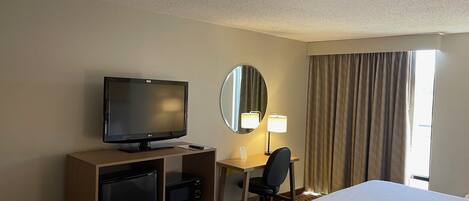 A hotel room with a bed, a desk with a computer, a television, a mirror, and a window with curtains.
