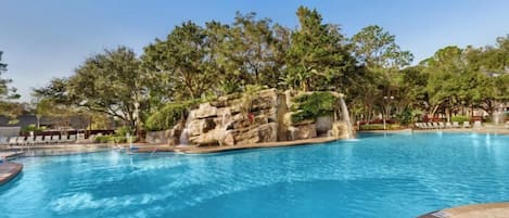 Una piscina con una cascada sobre rocas, rodeada de árboles y sillas de descanso.