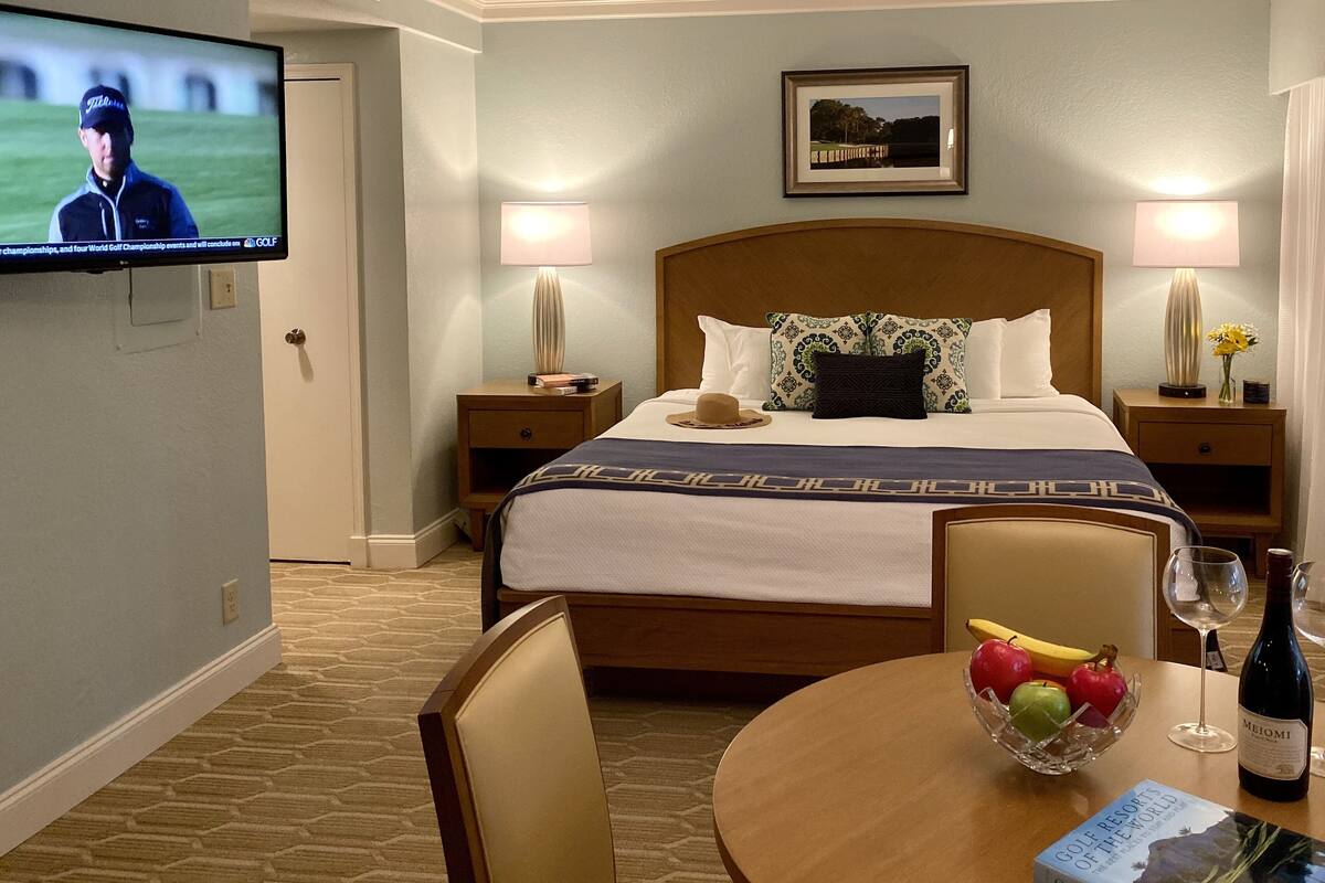 A hotel room with a bed, a TV, a dining table with fruit and wine, and a framed picture on the wall.