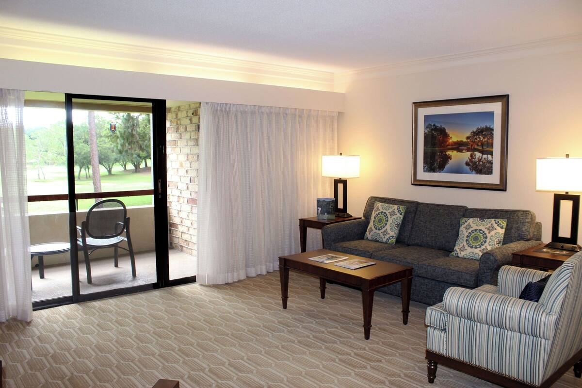 A living room with a sofa, armchair, coffee table, and two lamps. There is a window with a view of trees and a door leading to a balcony with a chair.
