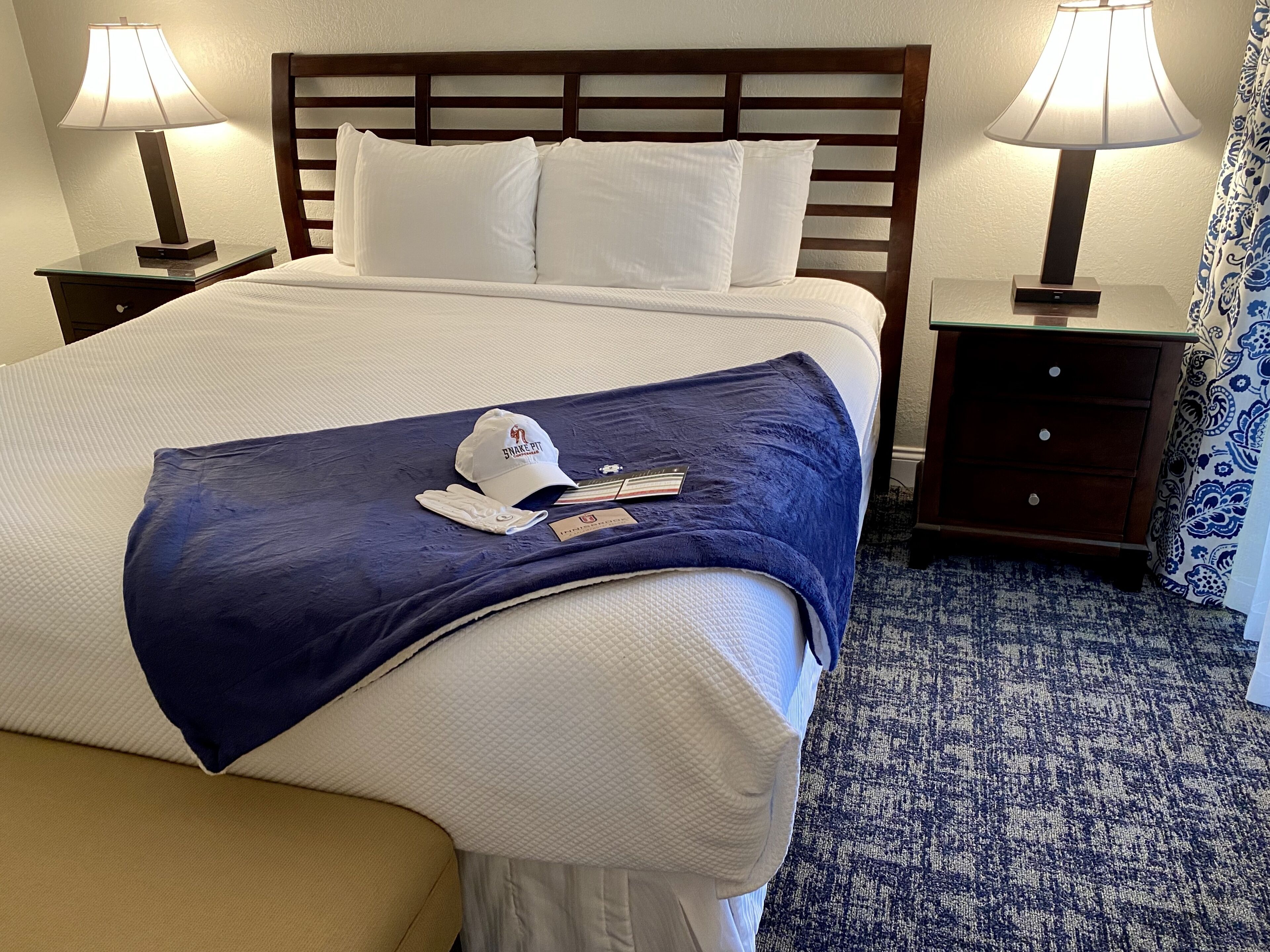 Un letto rifatto con cura, con una coperta blu, cuscini bianchi e un cappuccio bianco sul letto. Sono presenti due comodini con lampade, una testiera in legno e una tenda decorata.