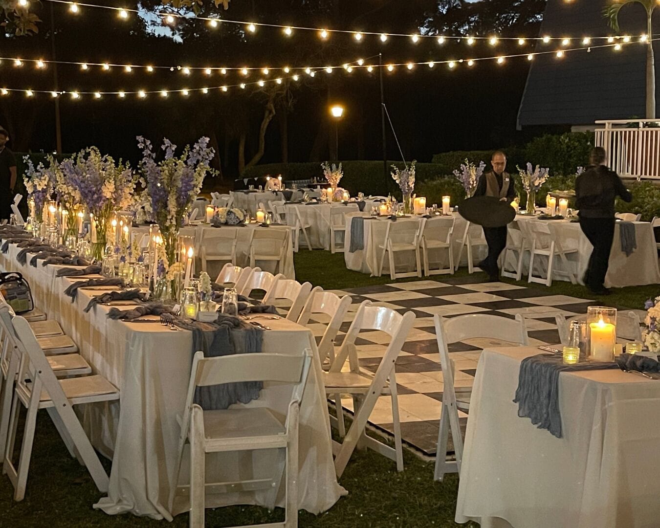 An outdoor event space with tables set for a formal gathering, decorated with floral arrangements and candles.