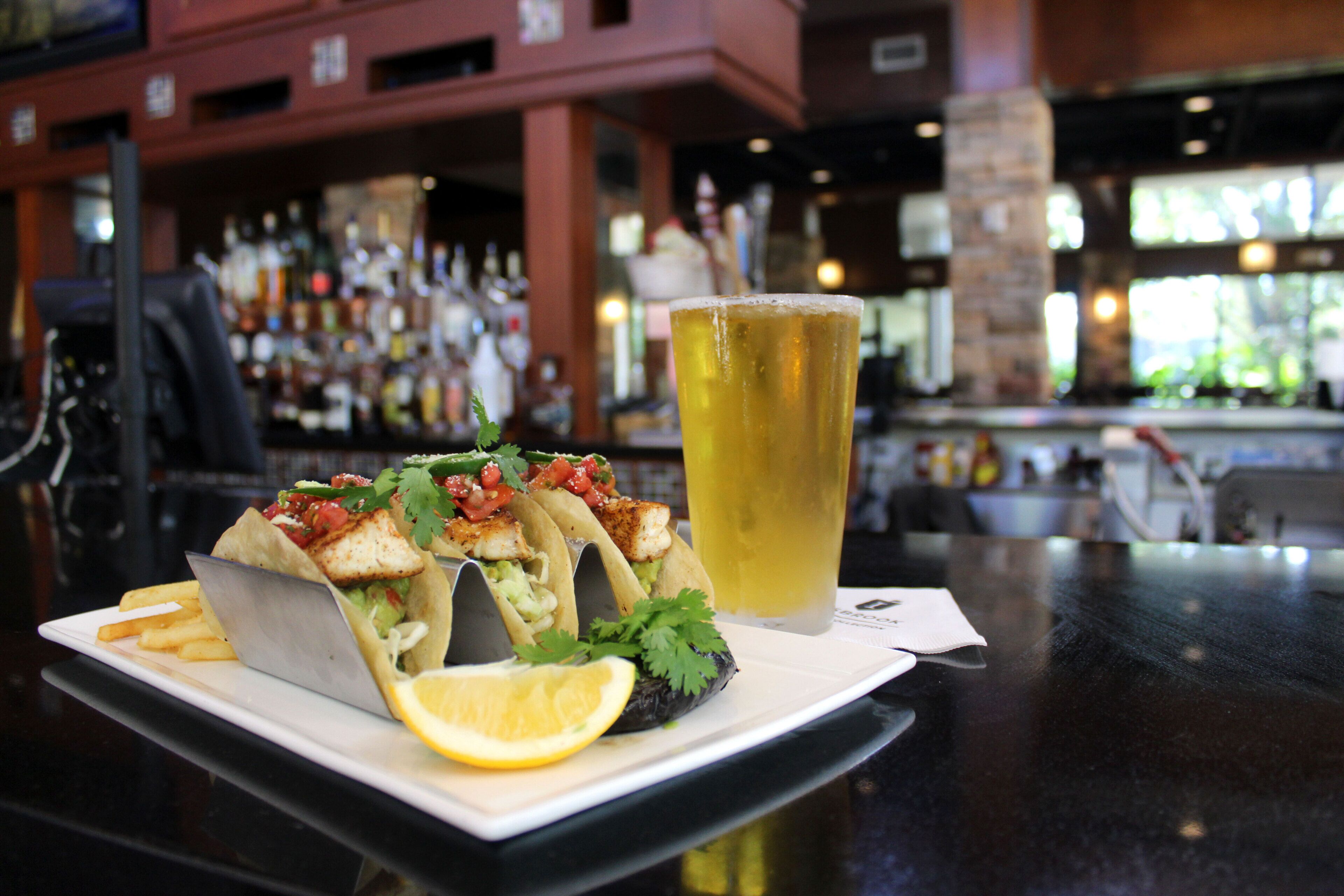 A plate with fish tacos, a glass of beer, and fries on a table in a bar setting.