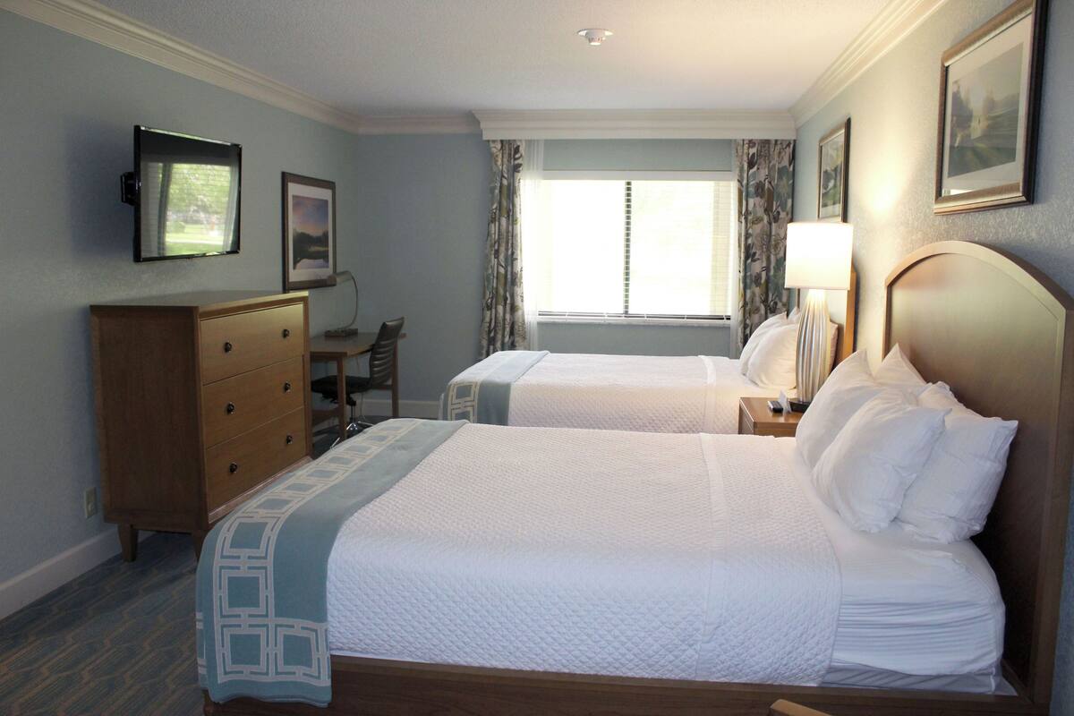 A hotel room with two beds, a wooden dresser, a desk, and a window with curtains.
