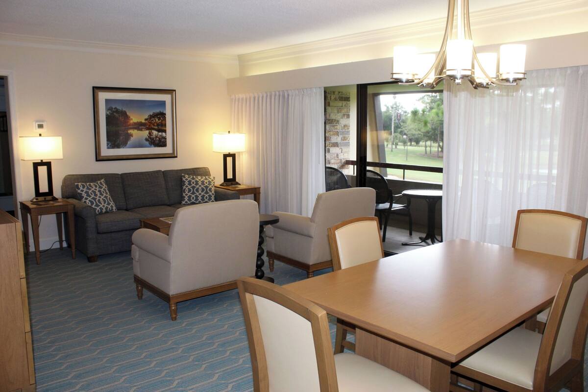 A hotel room with a dining area, a sofa, two lamps, a chandelier, and a window with curtains.